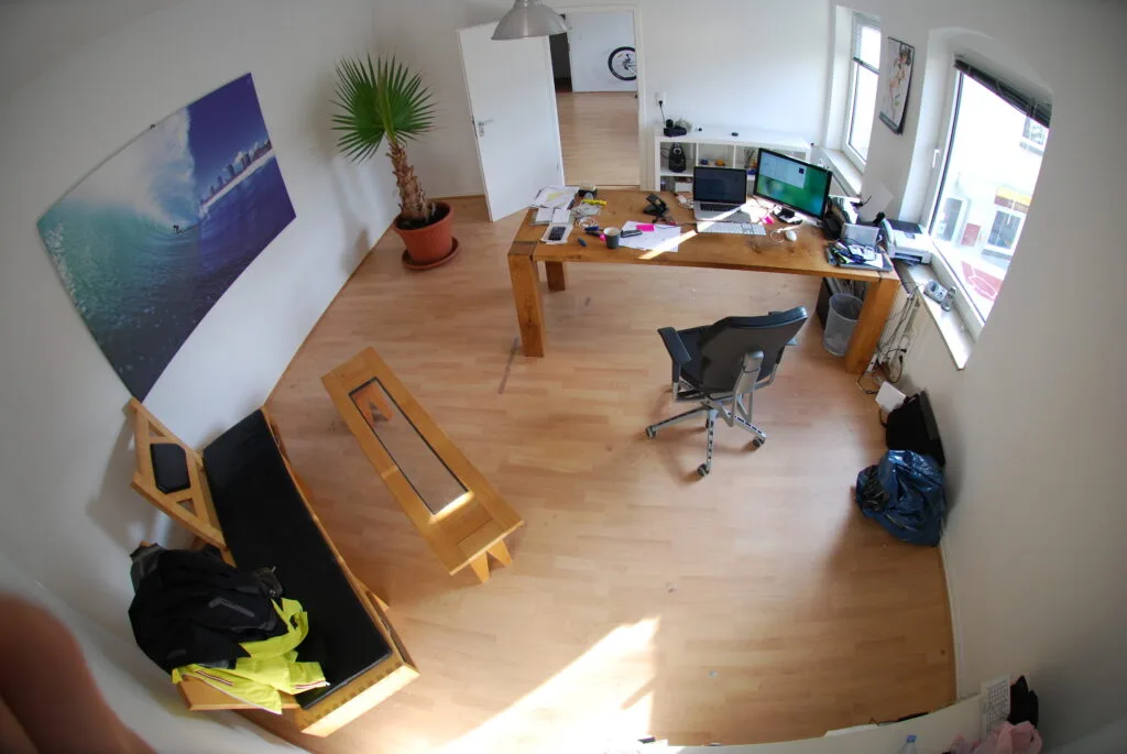 Foto mit Fisheye-Look aus der Ecke eines kleinen, leeren Büros mit weißen Wänden und hellem Laminatboden.