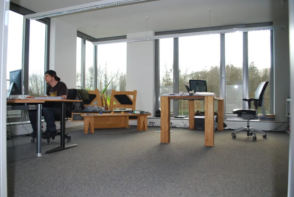 Das Innere eines Büroraums mit grauem Teppich und großen Fenstern. Eine Person sitzt an einem Schreibtisch und arbeitet.