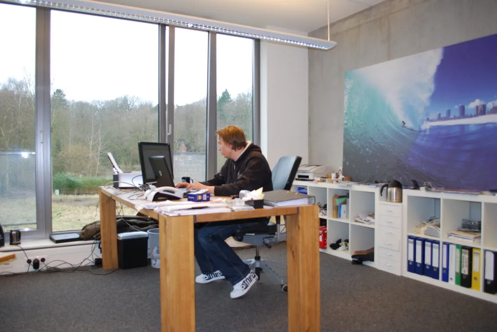 Kalle arbeitet an einem Schreibtisch in einem Büroraum mit grauem Teppich und großen Fenstern.