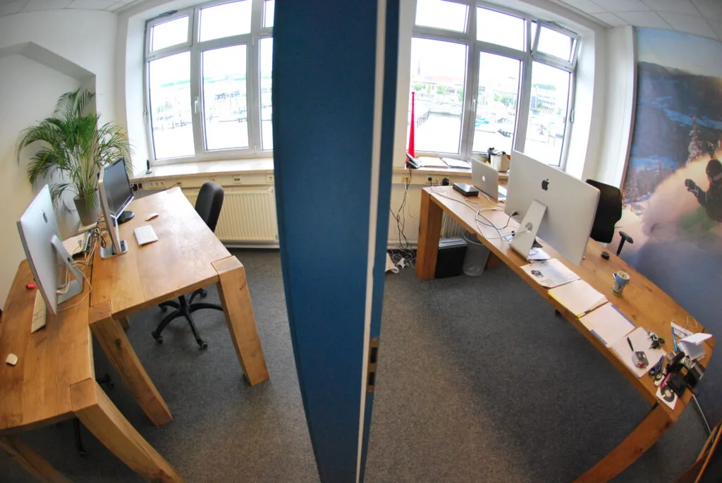 Foto zweier kleiner Büroräume mit drei Holzschreibtischen auf grauem Teppichboden.