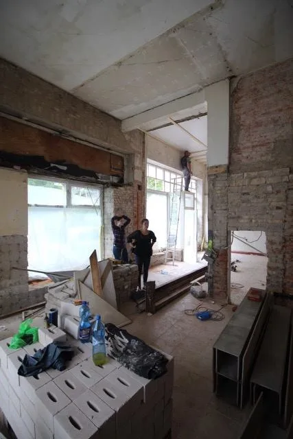 Foto eines in der Sanierung befindlichen Innenraums mit sehr hohen Decken. Drei Personen stehen inmitten der Baustelle.